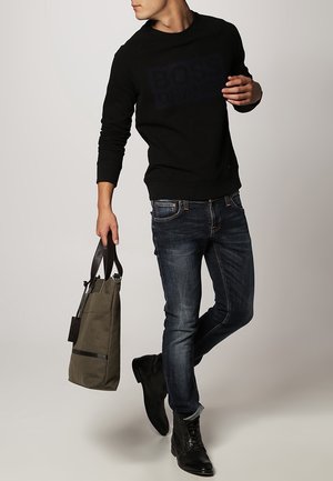 Felpa nera con logo testurizzato, jeans blu, stivali di pelle neri e borsa a tracolla in canvas verde oliva con strap a contrasto.