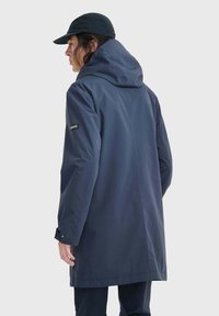 Manteau imperméable bleu marine avec capuche, arborant un logo sur la manche gauche. Texture lisse et longueur mi-cuisse.