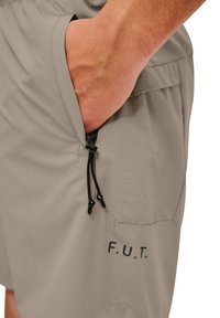 Beige shorts i lätt tyg med en rutmönstrad ficka, dragsko-stängning och tryckt "F.U.T."-logotyp på sidan.