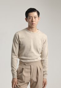 Suéter beige de manga larga con cuello redondo, combinado con pantalones de pliegues beige claros que presentan un detalle de botón en la cintura.