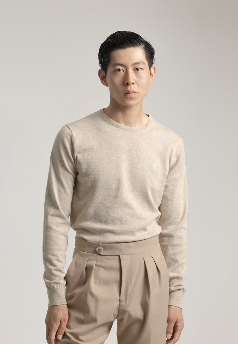 Suéter beige de manga larga con cuello redondo, combinado con pantalones de pliegues beige claros que presentan un detalle de botón en la cintura.