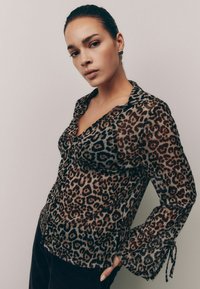 Prozračna bluza s leopard printom, s kopčanjem sprijeda, ovratnikom i lepršavim rukavima koji imaju vezice na manžetama. Smeđi i crni uzorak.