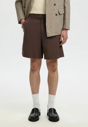 Henkilö, jolla on päällään ruskeat shortsit, beige bleiseri, valkoiset sukat ja mustat loafers-kengät, seisoo yhdellä kädellä taskussa yksivärisen taustan edessä.