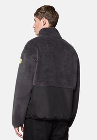 Polarowa kurtka w ciemnoszarym kolorze z czarną dolną częścią. Posiada wysoki kołnierz oraz mały, odblaskowy żółty akcent na ramieniu.