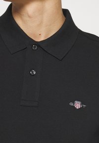 Homme portant un polo noir à col avec deux boutons et un petit logo GANT sur la poitrine.