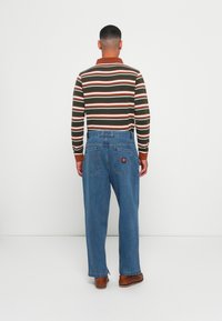 Gestreepte langemouwen shirt met bruine, groene en witte strepen, gecombineerd met een losse blauwe spijkerbroek voorzien van twee achterzakken en een logo patch.