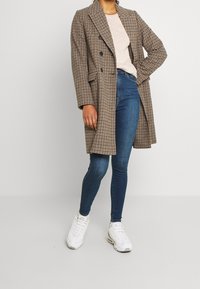 Mujer vestida con un abrigo marrón de cuadros, una blusa beige clara, jeans ajustados azul y zapatillas blancas, de pie con una mano en la cadera.