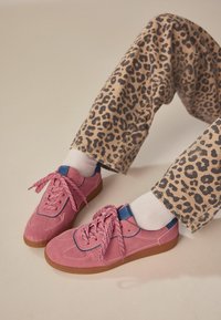Rosa mockasammet sneakers med blå detaljer och mönstrade snören, tillsammans med beige byxor med ett svart leopardmönster.