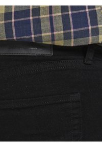 Mustat farkut, joissa on sileä pinta ja hienovarainen tikkaus yksityiskohtien lähellä taskuja, osittain peitetty sinisellä, keltaisella ja vaaleanpunaisella ruudullisella paidalla.