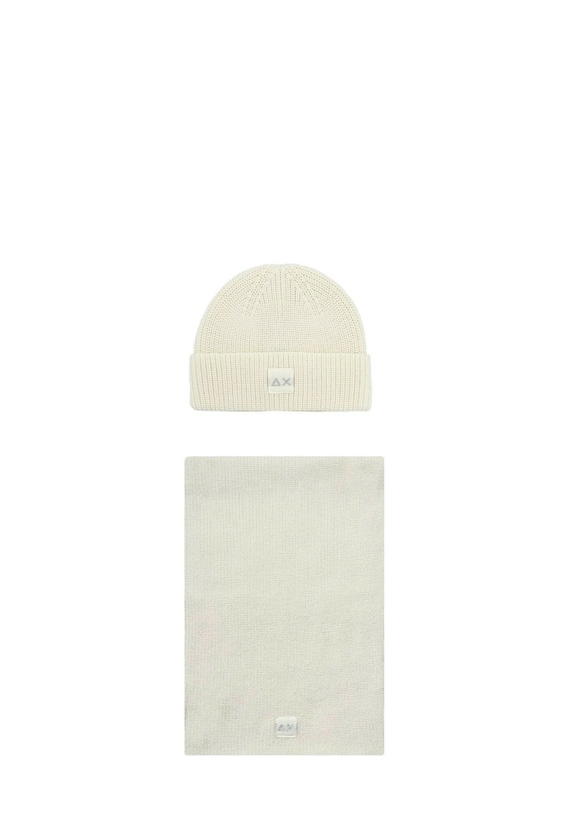 Berretto lavorato a maglia color crema e sciarpa rettangolare coordinata, entrambi con piccole etichette a logo quadrato, presentati su sfondo bianco.