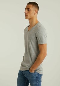 Tricou gri cu mânecă scurtă și decolteu în V, confecționat dintr-un material moale, cu o croială relaxată și detalii de cusătură simple, asortat cu blugi de culoare albastru închis.