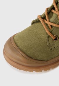Primo piano di una scarpa in pelle verde oliva con suola in gomma marrone e lacci marroni su sfondo bianco.