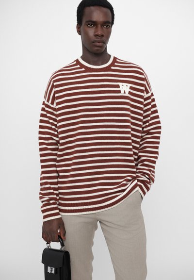 Suéter a rayas marrón y blanco con cuello de canalé. Presenta un logotipo blanco prominente en el pecho. La modelo sostiene un pequeño bolso negro.