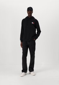 Schwarzer Hoodie mit Fronttasche, weiße Sneakers und weite schwarze Jeans. Der Hoodie hat ein kleines rot, weiß und blaues Logo auf der Brust.