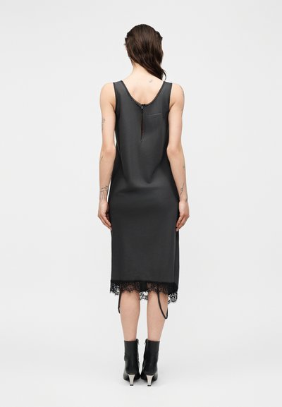 Γυναίκα που φοράει μαύρο μίντι φόρεμα χωρίς μανίκια με δαντέλα στο τελείωμα και σκίσιμο στην πλάτη, συνδυασμένο με μαύρα μποτάκια, στέκεται μπροστά σε απλό φόντο.