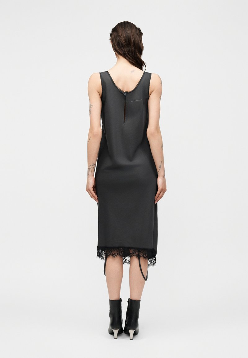 Frau trägt ärmelloses schwarzes Midikleid mit Spitzenabschluss und Rückenschlitz, kombiniert mit schwarzen Stiefeletten, steht vor neutralem Hintergrund.