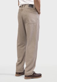 Pantalones de tiro ancho beige con una textura suave, bolsillos traseros y una etiqueta negra en la cintura. Combinados con calzado marrón oscuro.