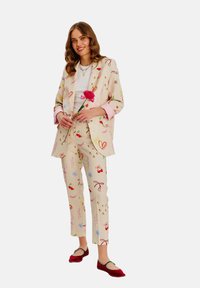 Beżowa marynarka i spodnie w wzory z kolorowymi rysunkami; biała bluzka; czerwone buty; trzyma różową kwiat. Miękka tkanina, luźny krój.
