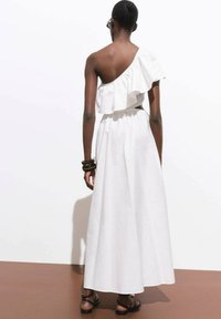 Robe blanche à une épaule avec un col à volants, taille élastique et jupe fluide. Portée avec des bracelets noirs et des sandales plates.