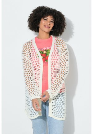 Vrouw met krullend haar die een witte opengewerkte cardigan draagt over een roze T-shirt met aardbeienprint en lichtblauwe spijkerbroek, glimlachend met gevouwen handen.