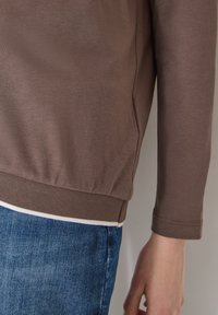 Bras droit et côté du torse portant une chemise marron à manches longues sur un jean bleu, sur un fond uni.