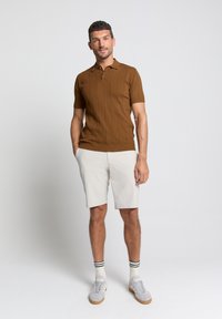 Bruine polo met korte mouwen en gestructureerde strepen, gecombineerd met lichtgrijze shorts. Model draagt witte sokken met groene strepen en grijze sneakers.