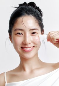 Una donna sorridente tiene un applicatore di gel traslucido contro la guancia. Ha la pelle liscia, capelli scuri e indossa una maglia bianca che lascia le spalle scoperte.