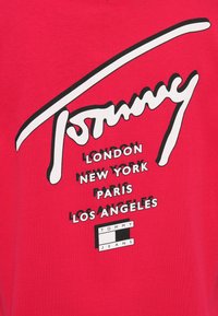 Červené bavlnené tričko s nápisom "Tommy" v kurzíve bielym písmom, pod ktorým sú názvy miest v čiernej farbe. Obsahuje logo "TOMMY JEANS" v čiernej krabici.