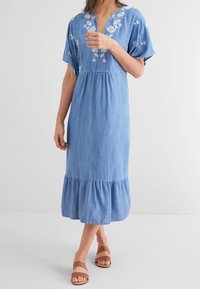 Blaues Chambray-Kleid mit kurzen Ärmeln, verziert mit floralem Stickereien am Ausschnitt und einem Rüschensaum. Kombiniert mit braunen Sandalen.