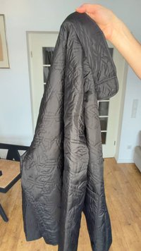 Ruka drži crnu prošivenu jaknu s geometrijskim uzorcima šavova u svijetloj sobi s drvenim podom i vratima s ostakljenim panelima.