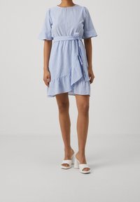 Robe rayée bleue avec manches courtes évasées, taille ceinturée et ourlet volanté. Associée à des mules blanches à talons et bouts ouverts.