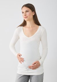 Langärmliges, V-Ausschnitt-Oberteil aus weichem weißen Stoff, figurbetonte Passform mit gerafftem Design an der Taille, kombiniert mit hellgrauen Jeans.