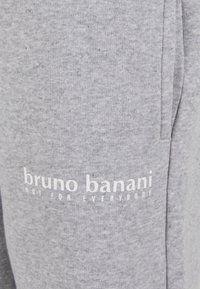 Pelēkas kokvilnas sporta bikses ar baltu logo un tekstu "bruno banani NOT FOR EVERYBODY" kreisajā pusē. Gluda tekstūra, ikdienišķs dizains.