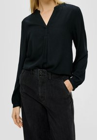 Blusa nera a maniche lunghe con scollo a V, caratterizzata da un leggero dettaglio in plissé frontale e tessuto testurizzato, abbinata a pantaloni scuri leggermente testurizzati.