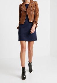 Kvinna iklädd en brun mocka motorcykeljacka över en marinblå knälång klänning med svarta ankelskor stående mot en vit bakgrund.