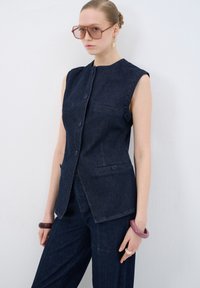 Chaleco sin mangas de denim oscuro con cierre de botones, dos bolsillos y una forma estructurada, acompañado de pantalones de pierna ancha a juego.