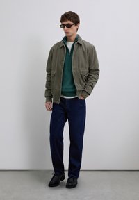 Un jeune homme portant une veste en velours côtelé olive, un pull à col zippé teal, un jean bleu foncé, des chaussures noires et des lunettes de soleil rectangulaires, se tenant à l'intérieur.