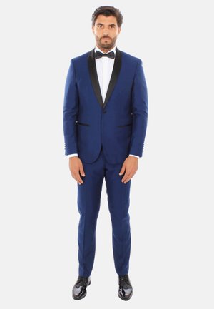 Tuxedo blu navy con rever neri, camicia bianca e papillon nero. Design aderente, texture elegante, con pantaloni slim-fit e scarpe nere lucide.