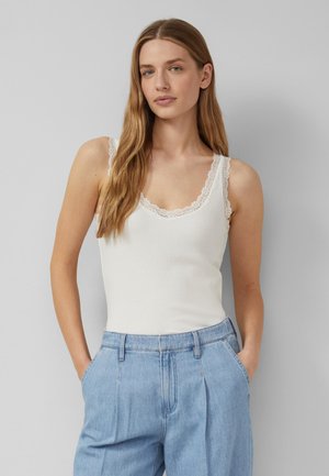 Kobieta z długimi jasnobrązowymi włosami, ubrana w biały bezrękawnik z koronką oraz jasnoniebieskie dżinsy z wysokim stanem, ręce w kieszeniach, neutralne tło.