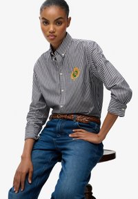 Stribet button-up skjorte i sort og hvid, med et broderet logo, parret med blå denim jeans og et brunt flettet bælte.