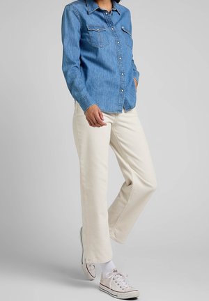 Person wearing a blue denim button-up shirt, off-white corduroy pants, white socks, and white low-top sneakers standing on one leg.