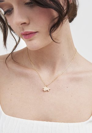 Tummahiuksinen nainen käyttää hienoa kultaketjukaulakorua, jossa on pieni siipinen sika-riipus, ja hänellä on päällään valkoinen teksturoitu toppi.