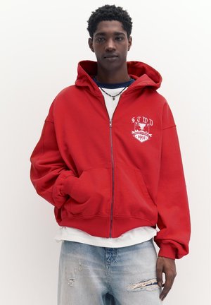 Mladić u crvenoj zip-up dukserici s bijelim grbom, slojevitim bijelim i tamnoplavim majicama te svijetloplavim poderanim trapericama, stoji na bijeloj pozadini.