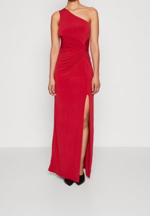 Vrouw die een rode, floor-length jurk met één schouder en een hoge split aan de dij draagt, met zwarte puntige schoenen, staand tegen een effen achtergrond.