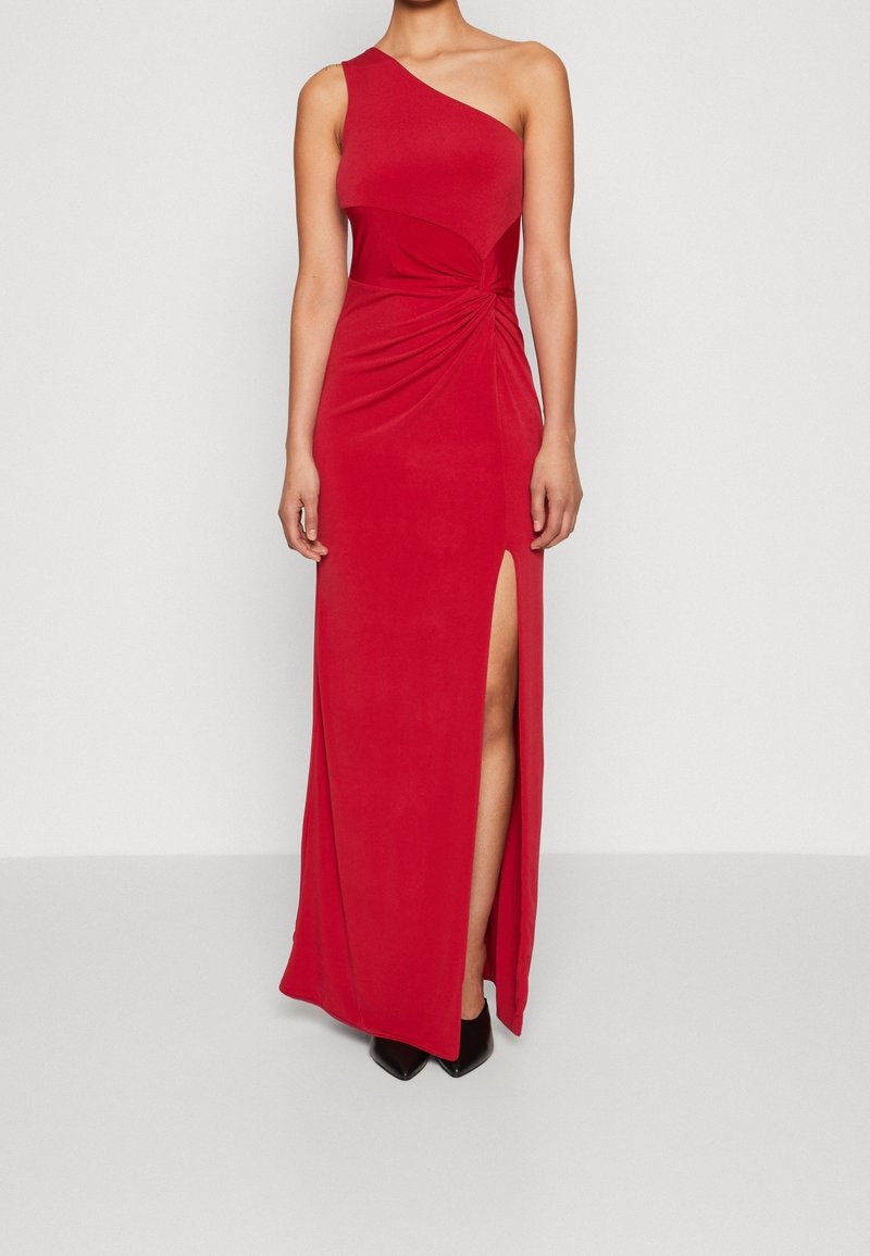 Femme portant une robe rouge longue jusqu'au sol, à une épaule avec une fente haute sur la cuisse et des chaussures noires à bouts pointus, debout devant un fond uni.