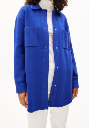 Person iført en knaldblå knapjakke med to store frontlommer over en hvid rullekrave og hvide bukser.