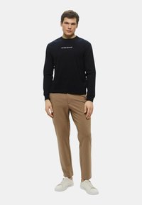 Maglietta nera a maniche lunghe con logo "GUESS JEANS", abbinata a pantaloni beige slim-fit e sneakers bianche. Tessuto liscio, design casual.