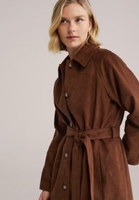 Trenci maro din piele intoarsă, cu rever decupat, nasturi mari și curea legată în talie. Are mâneci lungi și buzunare laterale.