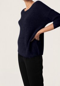 Marineblau gestrickter Pullover mit lockerer Passform, gerippter Textur und weitem Bootsausschnitt. Kombiniert mit schwarzen, eng anliegenden Hosen.