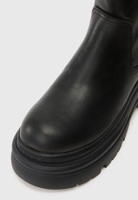 Bota de tornozelo em couro preto com biqueira arredondada, superfície texturizada, sola grossa e logo gravado na lateral. Design robusto e durável.